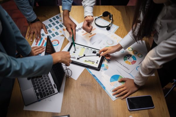 Four multi ethnic brokers analysing financial statistic on market using modern gadget at office. Male and female colleagues sharing with ideas and knowledge during briefing.